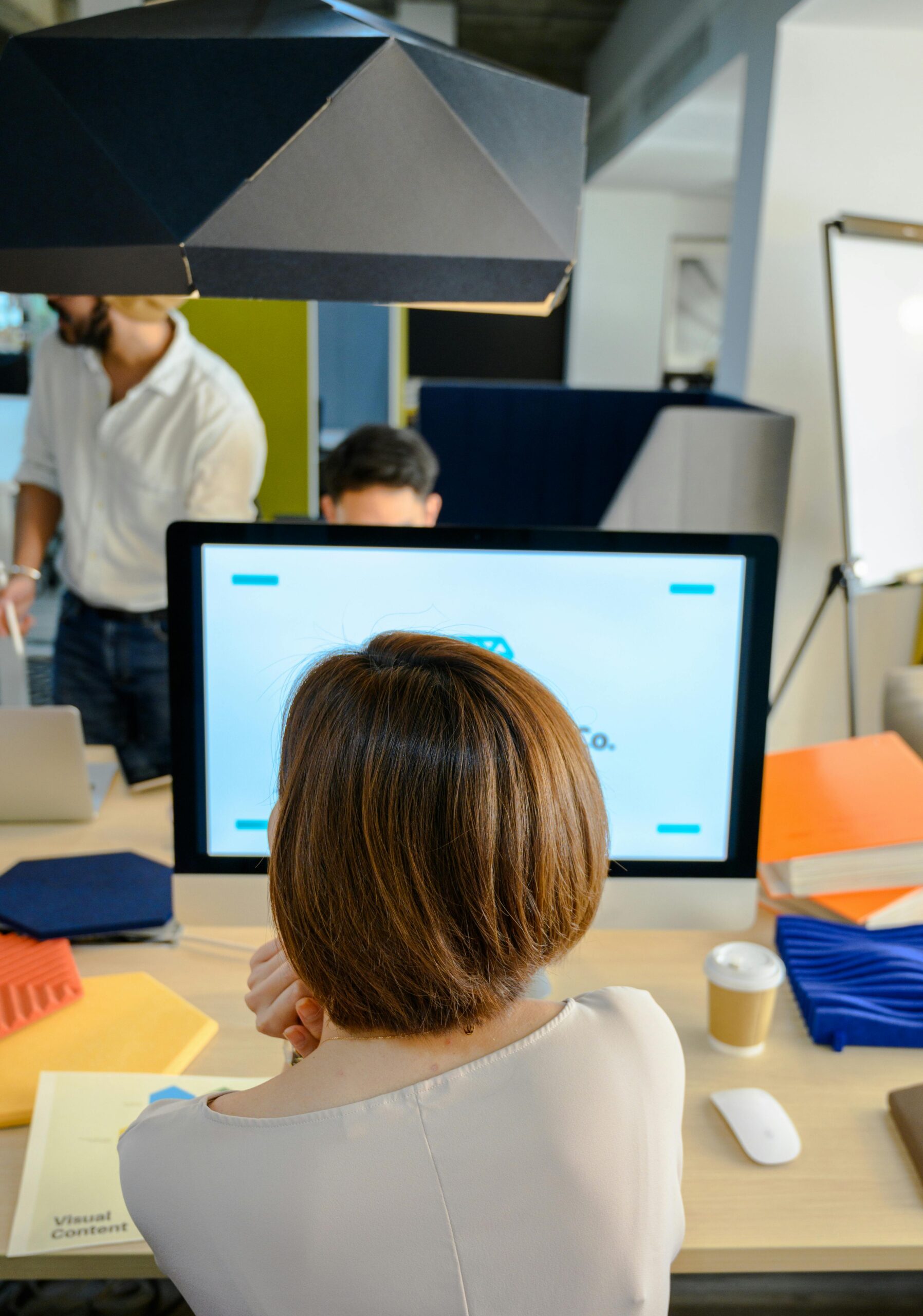 Woman working at a desk in a modern office with colleagues and computer monitors in view.